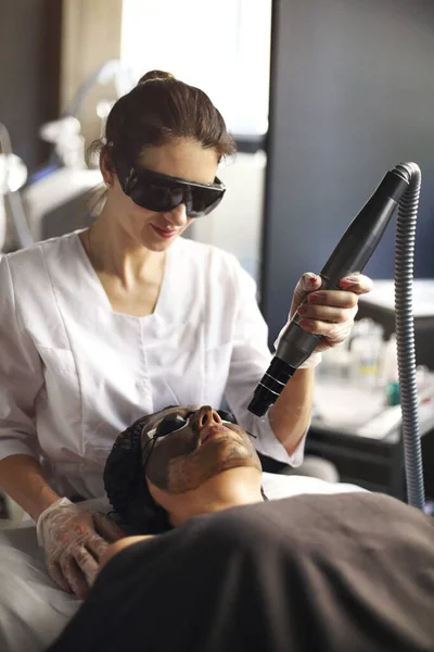 Beautician holding beauty laser near woman face covered with carbon mask and protective eyewear, spa procedures with laser and black masks