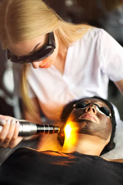 Beautician holding beauty laser near woman face covered with carbon mask and protective eyewear, spa procedures with laser and black masks