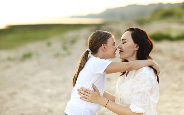 Loving mother kissing adorable girl in forehead while sitting together on shore in summer at sunset