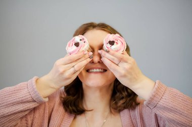 Zevkli bir kadın gözlerini lezzetli keklerle kaplıyor ve gri arka planda eğlenirken gülümsüyor.
