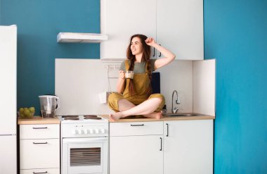 Genç çıplak ayaklı kadın tezgahta oturuyor ve modern mutfakta kahvenin tadını çıkarıyor.