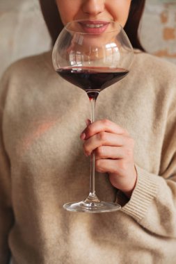 Kırmızı şarap kokan kazaklı mutlu bir kadın. Büyük bardakta, gri arka planda.