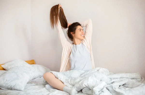 Jovem Encantada Com Cobertor Tocando Cabelo Sorrindo Enquanto Senta Cama — Fotografia de Stock