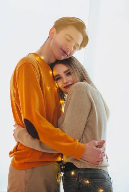 Gözleri kapalı genç bir adamın beyaz arka planda peri ışıklarıyla kız arkadaşını kucaklaması.