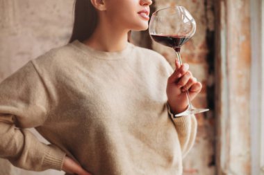 Kırmızı şarap kokan kazaklı mutlu bir kadın. Büyük bardakta, gri arka planda.