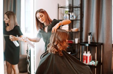 Young female hairdresser fixing hair of gorgeous smiling woman with hairspray. Hairdress and beauty concept