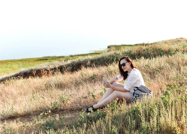 スマートフォンを持ちながら夏の時間に田舎の日当たりの良い芝生の上に座って モバイルで自分自身の写真を撮る美しい喜びのブルネットの女性 コミュニケーション アウトドア ライフスタイルリラックスコンセプト — ストック写真