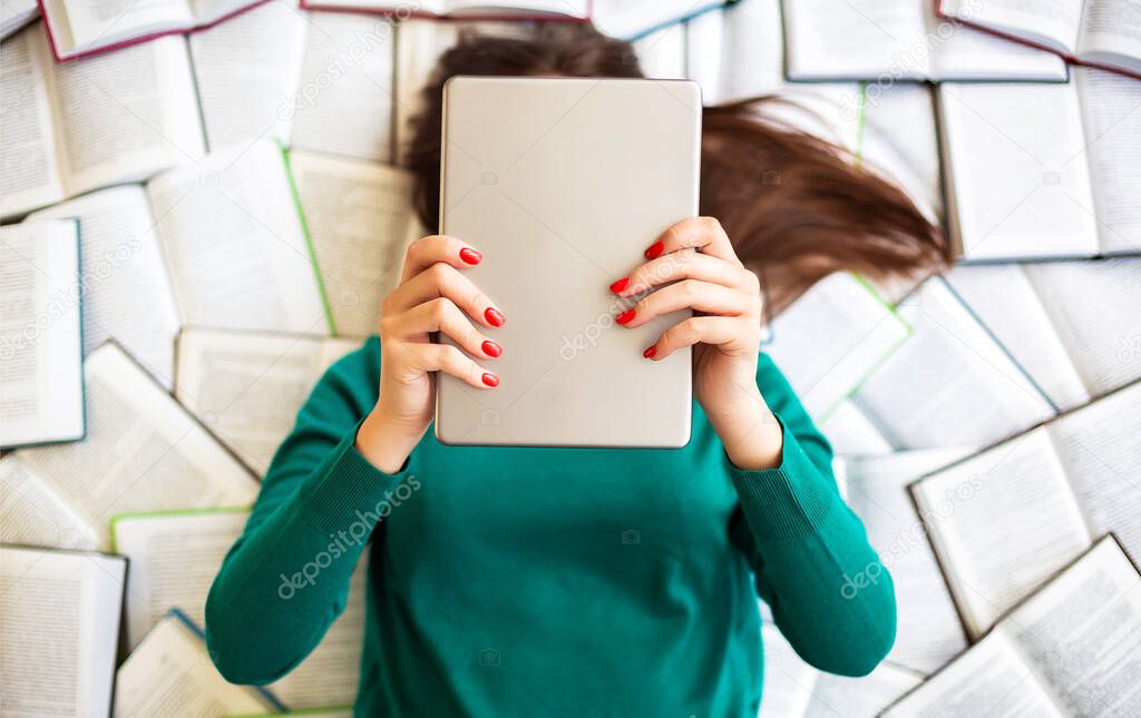 View from above of brunette woman in green sweater lies on large number of open books while holding digital tablet in hands in front of her face. Technology, educatio and communication concept