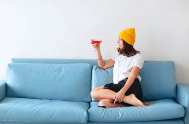 Elinde kırmızı alkollü içecekle elinde kokteyl bardağıyla evdeki kanepede oturan şık kıyafetler ve aksesuarlarla arkadaşlarıyla eğlenen genç bir kadının görüntüsü.