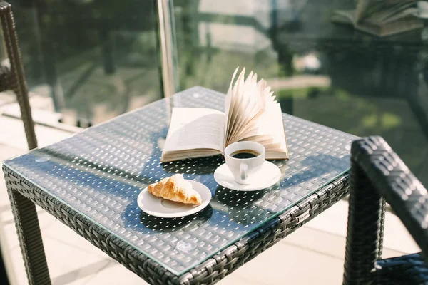 一杯黑咖啡 新鲜烘焙的羊角面包 在酒店阳台上的柳条藤桌上翻开的书 与模糊的绿色度假胜地景观相映成趣 暑假早上的早餐 — 图库照片