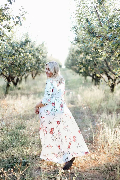 緑の梅の庭を歩いて長いロマンチックなドレスで幸せな若い屈託のない女性の垂直ショットは カメラで肩を上に見て 夏の日に自然の中で時間を過ごすと笑顔 — ストック写真