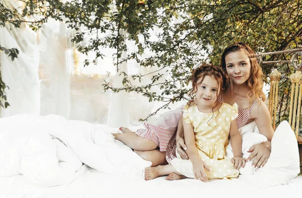 緑の庭できれいな白い寝具でベッドに座っている若い美しい幸せな家族 笑顔の母親と女の子の娘屋外で お母さんと子供は自然の中でリラックスし 夏の日を楽しんで — ストック写真