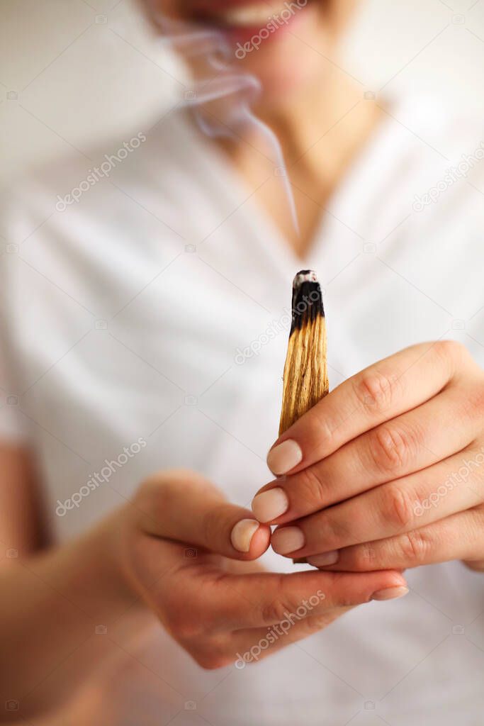 Atmospheric photo of smiling female SPA worker in white uniform holding burning palo santo incense stick with smoke slowly rising up. Spiritual practice, energy cleanse, healing, meditation concept