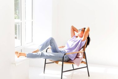 Uzun boylu, rahat Asyalı bir kadın. Gözleri kapalı, beyaz, boş bir odada, pencerenin önünde uzanıyor. Temiz havanın ve güneşin tadını çıkarıyor.