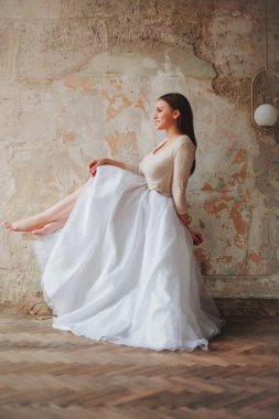 Güzel, genç, esmer, romantik bir kadın, uzun, tüylü, beyaz etekli, çıplak ayakla çatı katının bej odasındaki beton vintage duvara yaslanmış, yan bakıyor. Tam boy çekim