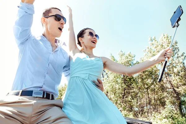 Young excited joyful couple, man and woman dressed festively making selfie with smartphone while sitting on car roof, screaming and smiling from happiness while raising hands up in air