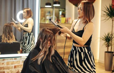 Mükemmel kıvırcık. Genç ve çekici bir kadın modern, aydınlık güzellik salonunda saçını yaptırıyor, profesyonel bir kadın kuaför saç maşasıyla saç ütülüyor ve şık bir saç kesimi yapıyor.
