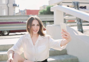 Güzel, genç, esmer kız şehir sokaklarında dolaşırken kendi fotoğrafını çekiyor. Dışarıdaki taş merdivenlerde oturuyor ve telefonu gözünün önünde tutuyor. Selfie çekiyor, gülümsüyor ve kamerada poz veriyor.