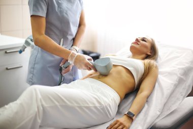 Güzelce gülümseyen sarışın kadın güzellik kliniğinde profesyonel cilt bakımı görüyor rahat sandalyeye uzanırken, doktor vücut şekillendirmek için özel ekipmanlar kullanıyor.