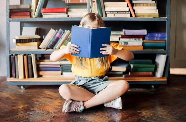 小女孩坐在家里的书架上 满屋子都是书 她的脸上挂着童话般的童话般的童话般的图画 孩子们喜欢读书 喜欢被新故事和新英雄环绕 — 图库照片