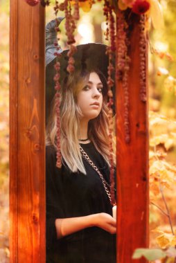 Cadı kostümlü ve şapkalı genç bir kadın, elinde mumla sonbahar ormanı arka planında izole bir şekilde Cadılar Bayramı fotoğraf çekimi için sahnelenmiş ahşap kapılar kullanıyor.