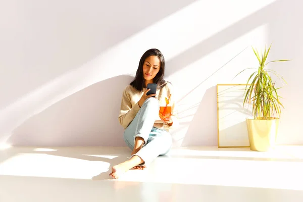 カジュアルな服を着た素敵な若いアジアの女性は フルーツカクテルを飲み 彼女の携帯電話でインターネットを閲覧し 自宅で日光浴部屋で床に座っている間に友人とオンラインでチャット — ストック写真