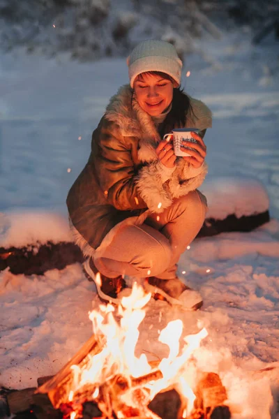 暖かいニット帽とジャケットに身を包んだ幸せな若い女性は 夜の雪で覆われた冬の森に囲まれた彼女の手に熱いお茶のカップとたき火で木製のそりに座っています — ストック写真