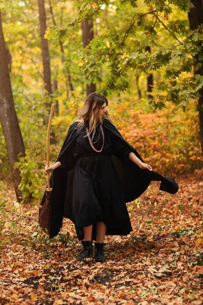 ブロンド女性で魔女の衣装でほうきで歩くと秋の葉で覆われたパス上のハロウィーンの日に森の中で — ストック写真