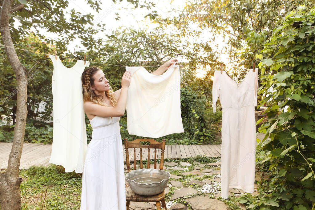 Young woman hanging laundry outdoors. Cute girl in dress washing white clothes in metal basin in backyard, hanging laundry on clothesline and leaving it to dry in garden, drying clothes outdoors in wind