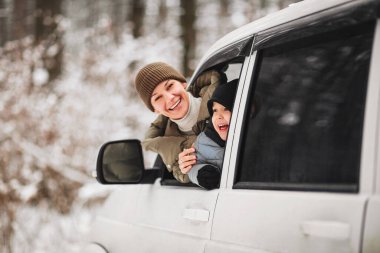 Neşeli yetişkin kadın ve dış giysili çocuk, kış ormanının bulanık arka planında araç camından dışarı bakarken kameraya bakıyorlar.
