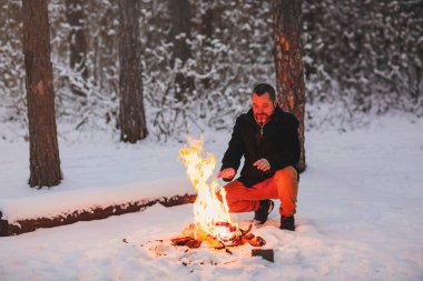 Olgun bir adam, soğuk kış akşamında kar ormanının ortasında kamp ateşi yaktıktan sonra ellerini ateşle ısıtarak kötü havalarda iyi ve rahat hissetmek, kış tatillerinin tadını çıkarmak için sıcağı kullanır.