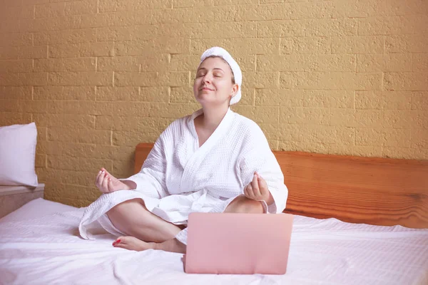 ローブのリラックスした女性は タオルはノートパソコンに耳を傾けハスの位置に瞑想します ホームスパリラクゼーション — ストック写真