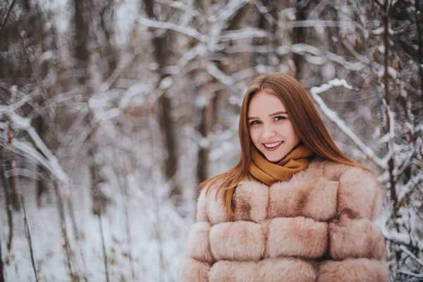 stock image Attractive young brunette woman wearing fur coat with scarf, standing alone in front of snowy forest, smiling at camera while spending time outdoors on winter day, selective focus on female