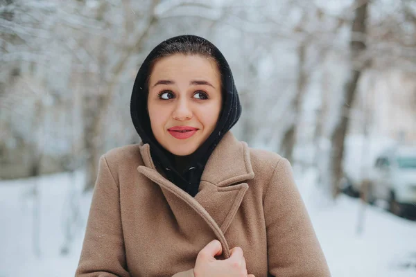 Young positive woman with funny smile on her face stands outside in beige coat and black scarf on head, shivering from winter cold weather against background of trees covered with thick layer of snow