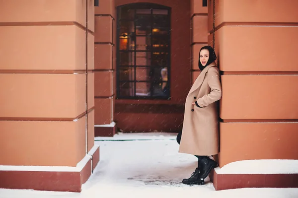 スタイリッシュなコートや革のブーツで若い美しい女の子孤独な建物の柱にもたれ外に立って 雪の冬の天気を楽しんで 最高の友人が来るのを待っている 人々のライフスタイルコンセプト — ストック写真