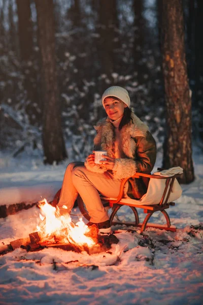 快乐的年轻女子头戴温暖的针织帽子 头戴紧身衣 心平气和地坐在篝火边的木制雪橇上 手里拿着一杯热茶 四周是白雪覆盖的冬季森林 — 图库照片