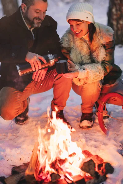 一緒に熱いお茶を飲む冬の雪の森の外のキャンプファイヤーで日光浴を愛する喜びの幸せな家族のカップルは 男と女は彼ら自身が抱き合って愛情を込めて見て暖かくなります トレッキング 季節の休暇 — ストック写真
