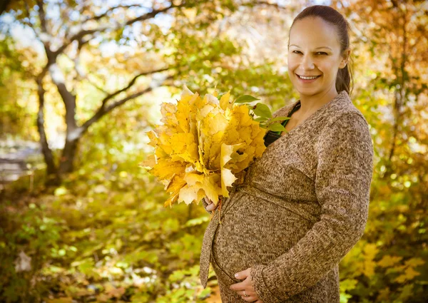 Woman holding hands with maple leaves on her pregnant belly — Stock Photo, Image