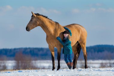 Genç ve tatlı bir kız, Trakehner buskin atını kışın arka planda kucaklıyor. Bir çocuk ve koca bir kısrak kışın bir tepede duruyor.