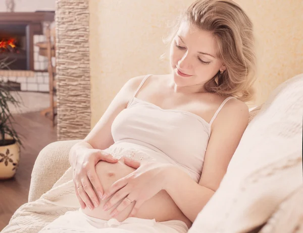 Feliz mujer embarazada sentada en el sofá y haciendo el gesto del corazón en — Foto de Stock