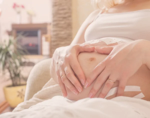 Mujer embarazada sentada en el sofá y haciendo un gesto de corazón en casa — Foto de Stock