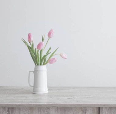 Beyaz seramik sürahide pembe laleler arka plandaki beyaz duvardaki ahşap masada.