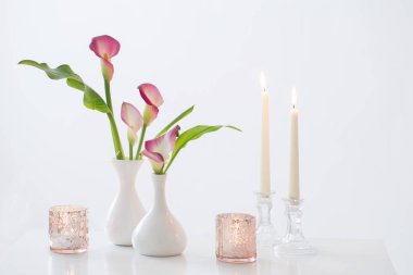 Vazoda pembe zambak ve beyaz arka planda yanan mumlar.