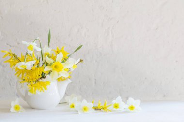 Beyaz arka plandaki ahşap masada vazoda beyaz ve sarı bahar çiçekleri.