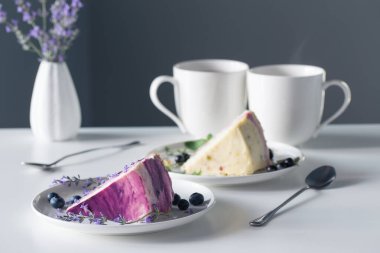 Böğürtlenli cheesecake ve arka plandaki gri duvardaki beyaz masada iki fincan çay.