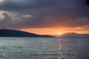 Taş plaj, güneş yolu, adaların siluetleri, gün batımı. Romantik deniz tatili. Sörfte kamera, dış hatlar
