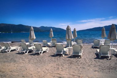 Akdeniz plajları turistleri, sahil kenarı tatil beldelerini, iklimsel sağlık merkezlerini bekliyor. Güneş pansiyonları, şezlonglar, şemsiyeler, yemek masaları ve sahildeki palmiye ağaçları.