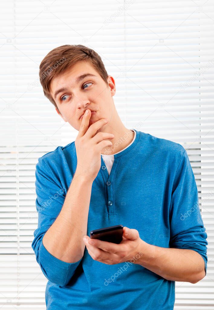 Pensive Young Man with a Mobile Phone is Thinking on the Jalousie Background in a Room