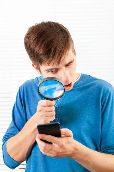 部屋で携帯電話を見て拡大鏡を持つ驚きの若い男 — ストック写真