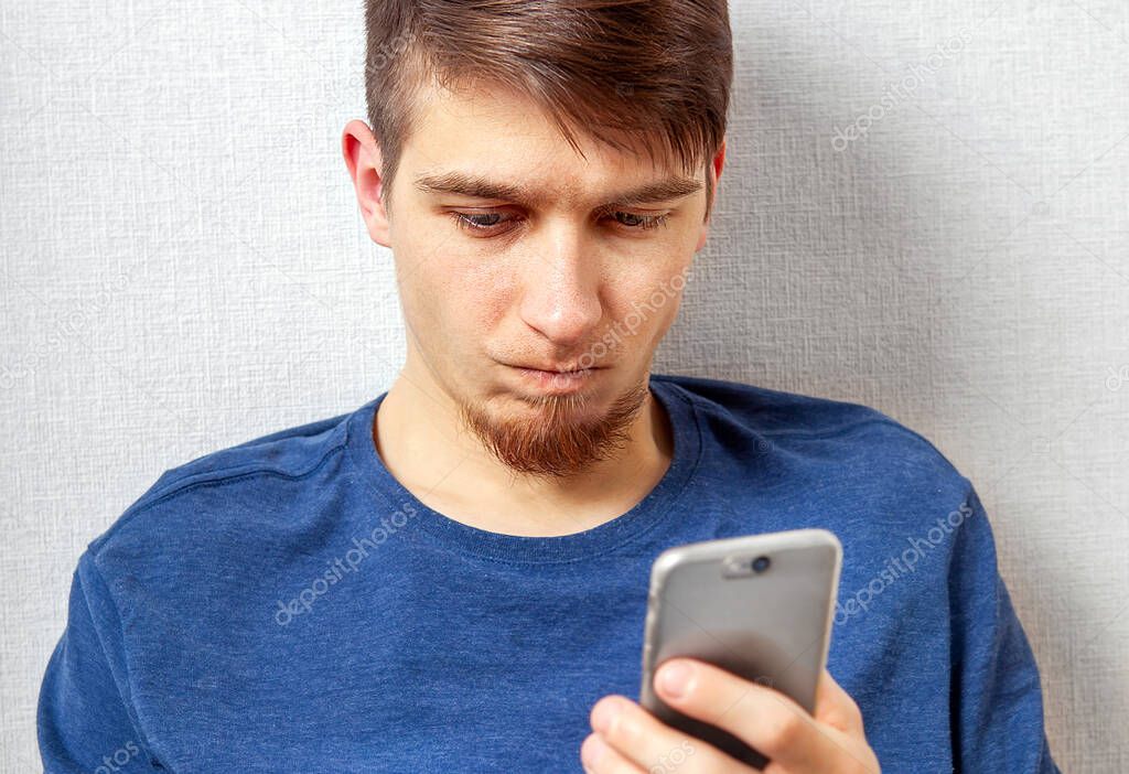 Dissatisfied Young Man with a Mobile Phone by the Wall in the Room